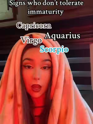 A selfie-style shot featuring a woman wrapped in a beige blanket, gazing directly at the camera with a slightly open mouth. The background is softly lit with red and brown hues. Overlaid text lists zodiac signs related to immaturity.
