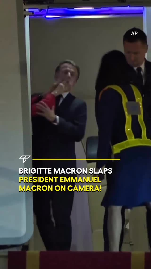 The cover image is a still from a video, capturing a moment in what appears to be a public setting. In the foreground, part of an individual, presumably a woman is shown with a yellow and black vest. Behind, Emanuel Macron, dressed in a dark suit, appears to be reacting to something. The background hints at the interior of a vehicle or a similar enclosed space. The text overlay states "Brigitte Macron Slaps President Emmanuel Macron On Camera!"