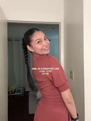 A woman with long braided hair is shown from the chest up, smiling and looking over her shoulder. She wears reddish-brown scrubs and stands in a well-lit hallway. Text reading 'DIML AS A PEDIATRIC CNA 4 AM - 8 PM ✨' is overlaid on the image.
