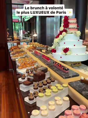 A well-lit shot of a luxury brunch spread. In the center, a multi-tiered white cake decorated with red and white flowers stands tall. Around the cake are multiple platters and displays filled with various pastries, desserts, and brunch items. The layout suggests a high-end dining experience, with a focus on presentation and variety.