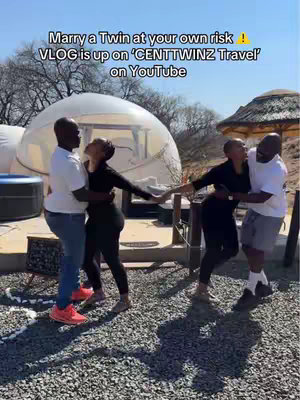The image shows two couples, presumably twins and their partners, embracing outdoors. The couples stand on a gravel surface with a blurred background featuring outdoor settings and a clear sky. Text overlays at the top state: "Marry a Twin at your own risk! VLOG is up on 'CENTTWINZ Travel' on YouTube".