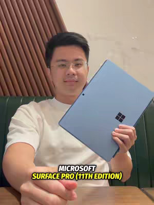 A man in a white t-shirt and glasses is smiling slightly, holding a light blue Microsoft Surface Pro tablet. The tablet faces the camera, partially obscuring his body. Behind him, a wooden slat design serves as a backdrop. Text overlays the image.