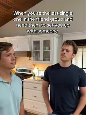 Two men stand in a kitchen setting, one looking off to the side, the other facing the camera with a contemplative expression. The background includes white cabinets and kitchen appliances.