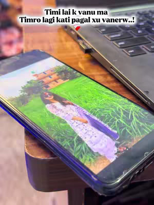 The image shows a smartphone displaying a photo of a woman in a long white dress and dupatta standing in a field of green grass with a building in the background. The phone sits at an angle, and above it is a laptop. Displayed above the phone is the text overlay in white font.