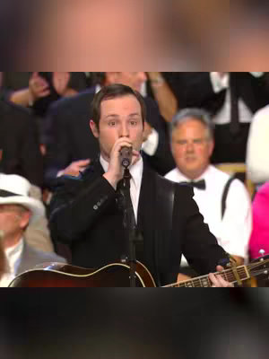 The image shows a man in a black suit and tie, holding a microphone and playing a guitar. He is positioned in front of a blurred background of other people, some of whom are also wearing formal attire. The composition is well-lit and creates a focus on the performer.