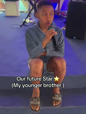 A young Black male sits on steps holding a microphone. Text reading "Our future Star (My younger brother )" is overlaid. Background is a stage setting.