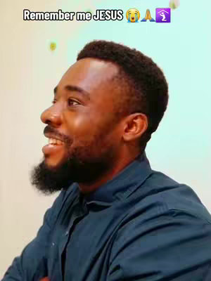 A man with dark skin and a trimmed beard is seen in profile, looking towards the left with a slight smile and open mouth. He is wearing a dark blue collared shirt. The background is a light greenish-blue color.