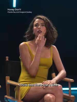 A woman in a yellow dress sits in a director's chair, holding her hand up to her face with a thoughtful expression. Text overlays indicate the title of a movie, 'Honey Don't!' and the names of the stars, Charlie Day and Margaret Qualley.