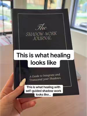 A person's hand holding a black book with the title 'The SHADOW-WORK JOURNAL' written on it. The hand has long, manicured nails. Overlayed on the book are two lines of white text that read 'This is what healing looks like' and 'This is what healing with self-guided shadow work looks like.' The background is blurred, but appears to be a light-colored room or outdoor space.
