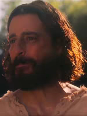A close-up shot of a man with a beard and long hair, lit from the side, with a soft focus. The man is looking off-camera with a slight expression of concern or contemplation.