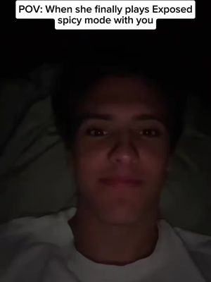 A close-up shot of a young man's face in a dimly lit room. He is gazing towards the camera with a slight smile, the lighting emphasizing his facial features. The background is dark and out of focus, suggesting the focus is solely on the man.