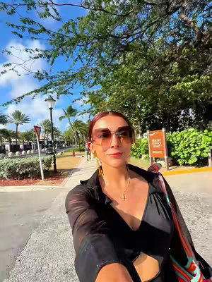 The image is a selfie with a woman wearing sunglasses, posing on a road. The background includes trees, buildings, and other people. The woman is the main focus, with the background providing context.