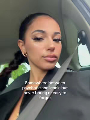 A young woman with long lashes, makeup, and a braid is captured from the chest up inside a car. The interior is visible; the seatbelt is in focus. Text at the bottom reads "Somewhere between psychotic and iconic but never boring or easy to forget".