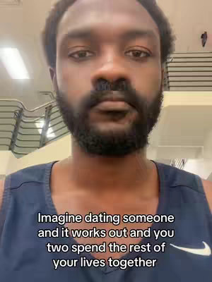 A close-up shot of a Black man looking directly at the camera. He has a short beard, and is wearing a navy blue tank top. Text overlayed on the image reads, “Imagine dating someone and it works out and you two spend the rest of your lives together”. The background appears to be a gym or an indoor space with metal railings and a bright light.