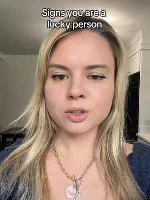 A woman with long blonde hair, a gold necklace, and dark eyeliner looks directly at the camera against the backdrop of a room with white walls and visible kitchen cabinets. Text overlayed at the top says, 'Signs you are a lucky person.'