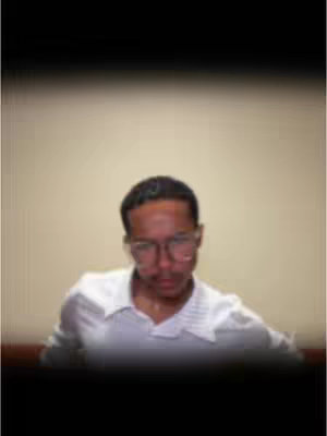 A person with short dark hair wearing glasses and a white collared shirt. The background is a light beige color, with a dark blurred band at the bottom and top of the image. The person is looking down.
