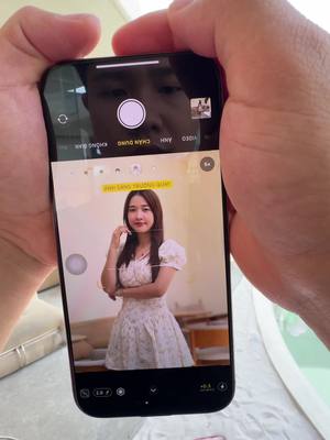 A person is holding an iPhone displaying a portrait of a woman. The phone's camera interface is visible, with the woman in focus. The phone is held in front of the person taking the picture to capture the image on the screen.