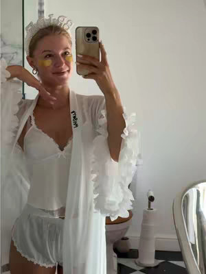 A woman in a bridal outfit takes a selfie in a bathroom setting. She wears a white robe with ruffled sleeves over a white lingerie set, and a 'Bride' headband. Gold eye patches are visible. The bathroom has a black and white checkered floor and a toilet in the background.