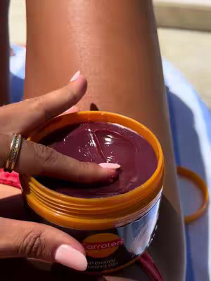 A close-up shot shows the arm and thigh of a person sunbathing. The focus is on a container of tanning gel; a finger is pressed into the gel, revealing a deep, reddish-purple color. The hand holding the container has perfectly manicured, light pink nails. The container is a bright orange color with a black label.