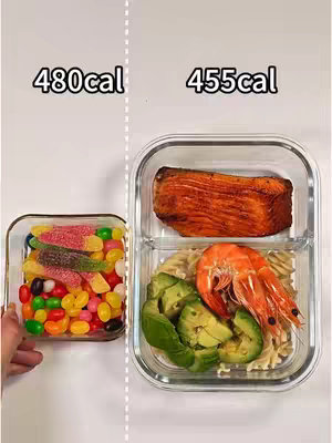 A split-screen image displaying two different food arrangements inside clear containers labeled with calorie counts. The left side shows colorful candies and jelly beans, labeled as 480 calories, inside a small square glass container. The right side features a meal of a grilled salmon fillet, shrimp, avocado slices, pasta on top of a white background, labeled as 455 calories, displayed in a larger divided container. The white background enhances the visibility of the food and calorie counts.