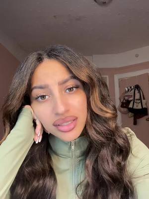 The image is a selfie of a young woman with long, wavy brown hair and a light green zip-up top. She is looking at the camera with a slight smile and her left hand is resting on her cheek. The background is out of focus, revealing a room with a ceiling light and part of a door with coats hanging on it.