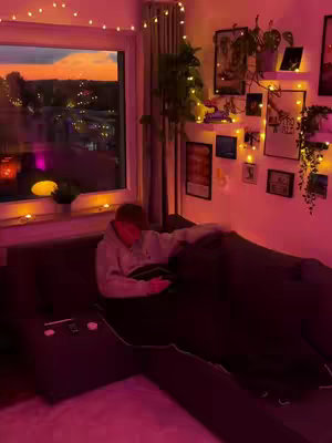 A warmly lit room featuring a window overlooking city lights, a couch with a person, surrounded by decorative lights and plant life.