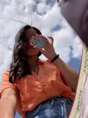 A low-angle shot captures a young woman from the chest up against a backdrop of a blue sky dotted with white clouds. She is holding a small card up in front of her face, partially obscuring it, with the card's design visible. The woman's arm is visible with a beaded bracelet. She's wearing an orange button-up shirt and blue jeans. The composition is framed as if the viewer is looking up at her.
