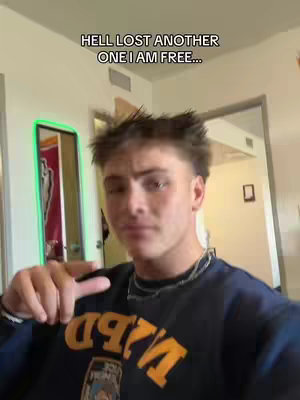 A young man in a dark blue sweatshirt with an orange logo is the primary focus in a selfie-style shot. The background is a softly lit interior. Above the subject's head, white text states, "HELL LOST ANOTHER ONE I AM FREE..."