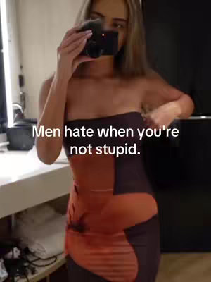 A woman is in a well-lit room, holding a digital camera in front of her face, partially obscuring it. She is wearing a strapless patterned dress. Text is overlaid on the image.