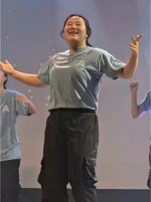 A person with arms outstretched, smiling, on a stage with a light blue background. There are other people on stage wearing similar shirts.