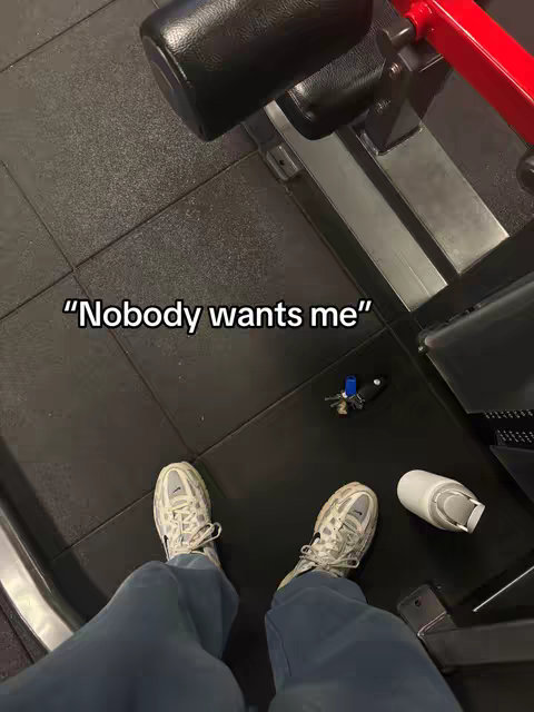 A first-person perspective shot of a person wearing sneakers and blue sweatpants in a gym. Overlayed text reads "Nobody wants me". The person's feet are in the foreground with gym equipment visible in the background, along with a water bottle and keys.