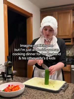 A person wearing a white bonnet and a dark, modest outfit stands in a kitchen, holding a knife and a cucumber, in front of a cutting board. The kitchen has wooden cabinets. A bowl of cut vegetables sits in the foreground, and an oil bottle.