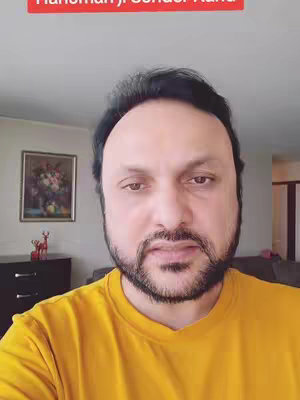 A man with a beard and a yellow shirt is filmed from the chest up. Red text appears at the top above his head. Behind him is a picture on the wall and a cabinet and deer sculpture.