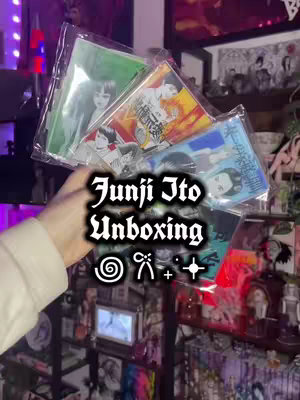 A hand holds several translucent plastic sleeves containing images from Junji Ito manga. The text 'Junji Ito Unboxing' and decorative symbols are overlaid on the image. The background displays a collection of manga, collectibles, and furniture.