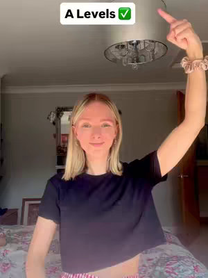 A young woman with blonde hair stands in a bedroom, pointing upwards with her right arm. A text overlay reads 'A Levels ✓.' The background includes a bed, a mirror, and a ceiling light.