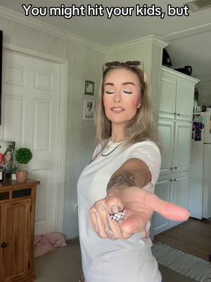 A woman with blonde hair stands in a well-lit room, holding her arm out towards the viewer. The text overlay, 'You might hit your kids, but...' is at the top, with the woman's face in focus, revealing a slight smile and closed eyes. She wears a white t-shirt and casual clothing with some visible tattoos on her arm, with sunglasses on top of her head, and a ring on her finger. The background highlights a part of an interior setting, with a view of a white door, cabinet, and a framed picture and some plants.
