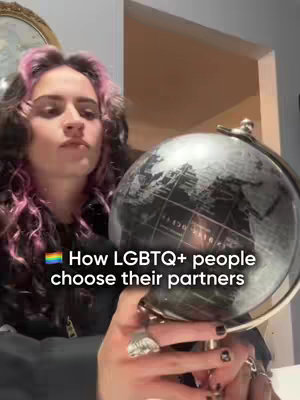 A woman with pink-streaked, dark curly hair looks up slightly, holding a globe in front of her. Text overlayed on the image states "How LGBTQ+ people choose their partners". The globe is dark, with a silver stand and detailed markings. The scene is lit with daylight, suggesting an indoor setting. The woman is wearing rings.