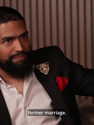 A medium shot featuring a Black man wearing a black blazer with a jewel-encrusted leopard pin and a red pocket square. He's wearing a white undershirt and a gold necklace. His expression is contemplative, with a subtle hint of a smile, suggesting he is listening or reflecting. The background has vertical stripe pattern.