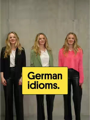 Three women stand in front of a gray background. A yellow rectangle is in the foreground with the text "German idioms." The women are wearing different colored tops. The woman on the left is wearing a black blazer, a white shirt, and black pants. The woman in the middle is wearing a green blazer and a white shirt with black pants. The woman on the right is wearing a pink sweater with black pants.