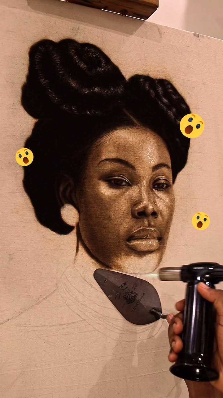 A close-up studio shot featuring a detailed charcoal drawing of a Black woman's face. The artwork is on a light-colored canvas with visible pencil guidelines. The woman is depicted with dark hair styled in an intricate updo. The artist's hand, holding a torch, is visible near the bottom right, near the work of art, and the torch light is hitting the bottom of the canvas. The overall effect is a blend of artistry