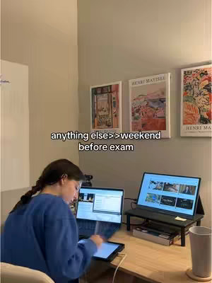 A person is sitting at a desk with two laptops and a tablet displaying content. There are also some posters on the wall behind the desk. The person appears to be wearing a blue sweater and has their hair in a braid. The scene is well-lit and has a clean, organized feel to it.