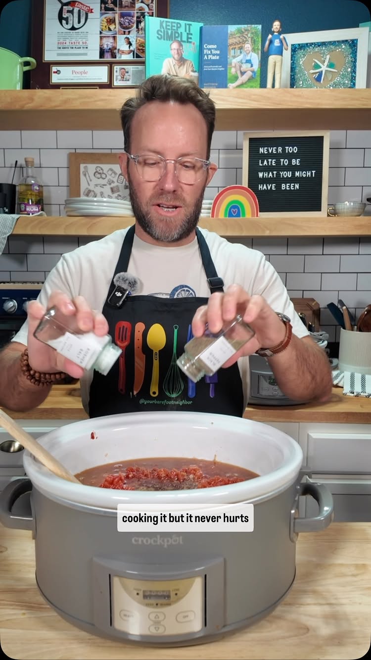 A man in a kitchen is pouring spices into a slow cooker filled with food.  He is wearing an apron, and there are shelves and cookbooks in the background. The text overlay reads, 'cooking it but it never hurts.'