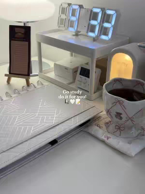 An aesthetically pleasing desk setup featuring a white desk, a digital clock, notebooks, a cup of tea, and other study-related items. The image is well-lit, suggesting a calm and inviting study environment.