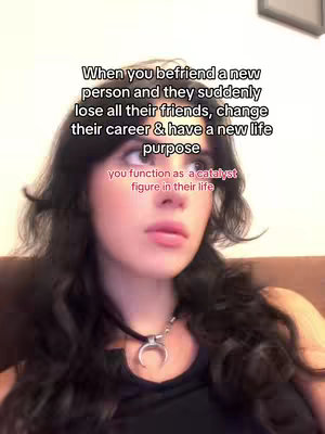 A young woman is centered in a selfie-style shot. She has long, dark wavy hair and she's looking off to the side of the camera, appearing contemplative. The text overlay features white text on a blurred background that reads 'When you befriend a new person and they suddenly lose all their friends, change their career & have a new life purpose'. The bottom text reads 'you function as a catalyst figure in their life'. She wears a black choker with a crescent moon pendant. The overall lighting is soft, and the palette is muted.