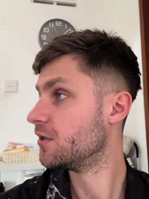 A man with short, dark hair, stubble, and a black shirt is seen in profile. The background includes a white wall with an analog clock and a blurry door frame.