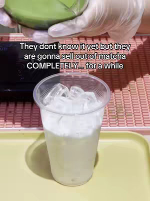 The image shows a clear plastic cup filled with ice and a milky liquid, likely a matcha latte. A hand wearing a white glove holds a pitcher of matcha above the cup, about to pour green matcha. The background is a light pink surface, and there is text overlayed across the top of the image.