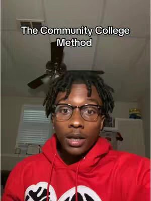 A young man with short, dark hair and glasses wearing a red hoodie addresses the camera. The background includes a ceiling fan and part of a window.