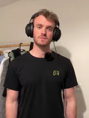 A man with light skin and short brown hair is centered in the frame, wearing a black t-shirt and large black headphones. A small microphone is clipped to his collar. Behind him is a blurry interior with elements suggesting a wardrobe and a plain wall. The lighting is soft, and and he looks directly to the camera.