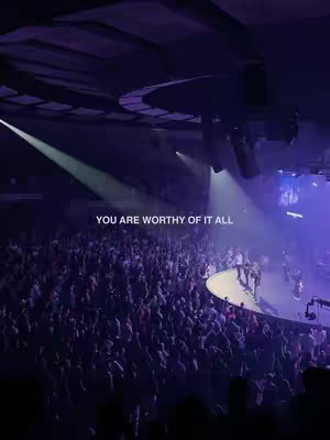 A wide shot of a large auditorium filled with people, likely a church service. Stage lights illuminate a stage where a band is positioned, with a large crowd facing them. The overall color scheme is a mix of dark and light purples, with bright spotlights emphasizing the stage.
