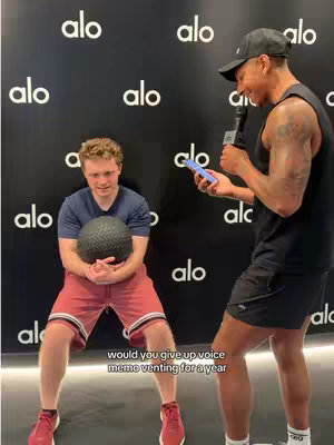 Two men are in a fitness studio with a black backdrop that has the brand 'alo' printed all over it. A man on the right is holding a phone and a microphone, seemingly interviewing another man who is holding a medicine ball. Text on the bottom is in white.